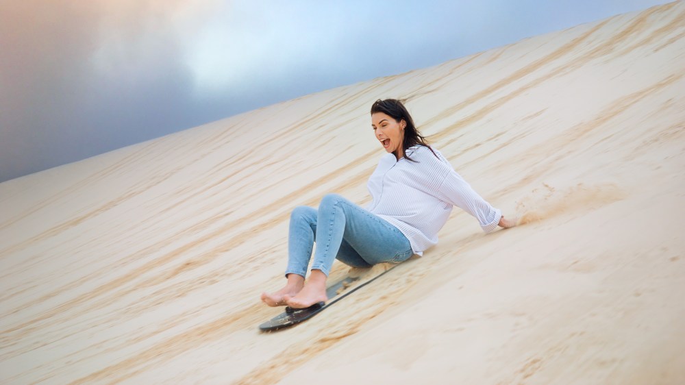 Person sliding down a sand dune on a board, wearing a white shirt and jeans.