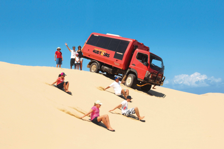People sandboarding down a dune near a red 4WD tour truck under a clear blue sky.