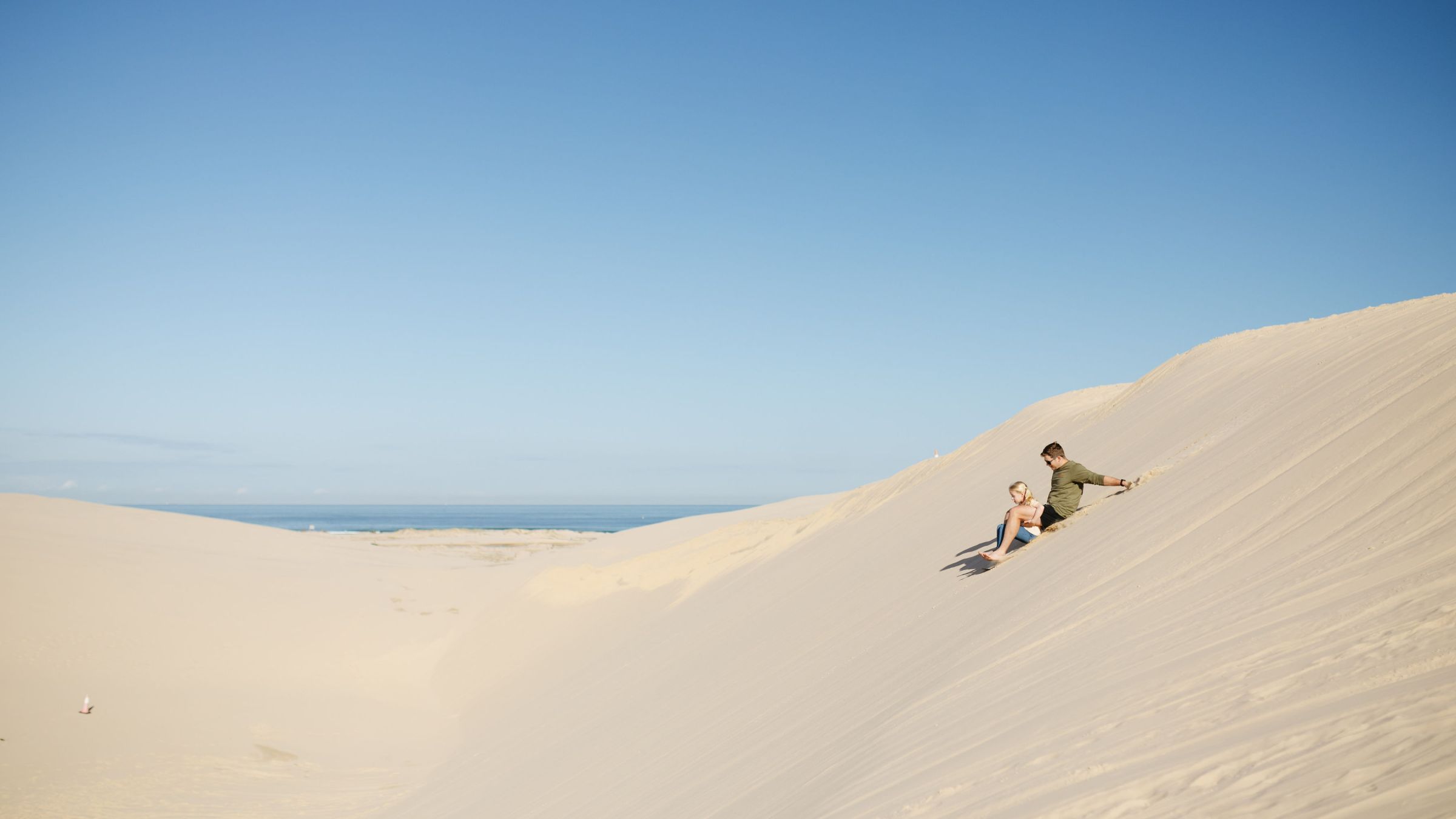 Two people sliding down a sand dune under a clear blue sky.
