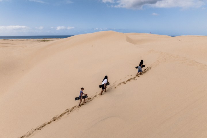 Three people walk up a sand dune carrying boards under a blue sky.
