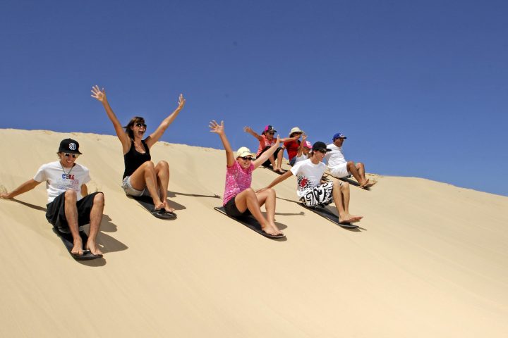 Group of people sandboarding down a dune under a clear blue sky.