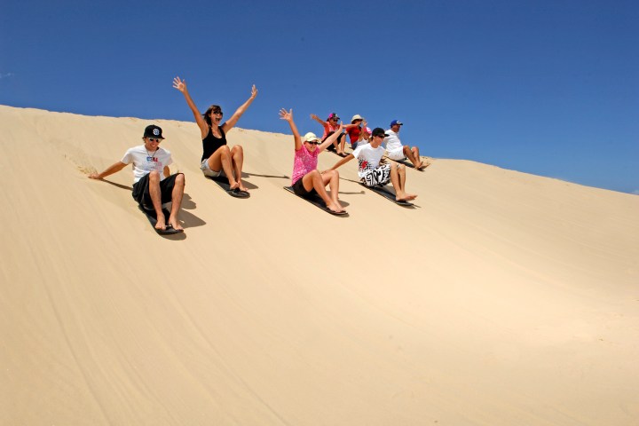 Group of people sandboarding down a dune under a clear blue sky.