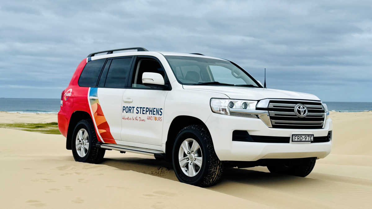 White SUV with colorful decals driving on sandy beach under cloudy sky.