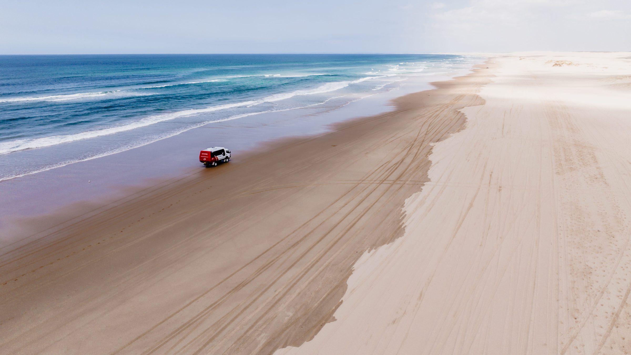 A red and white truck driving on a wide sandy beach near the ocean with blue skies.