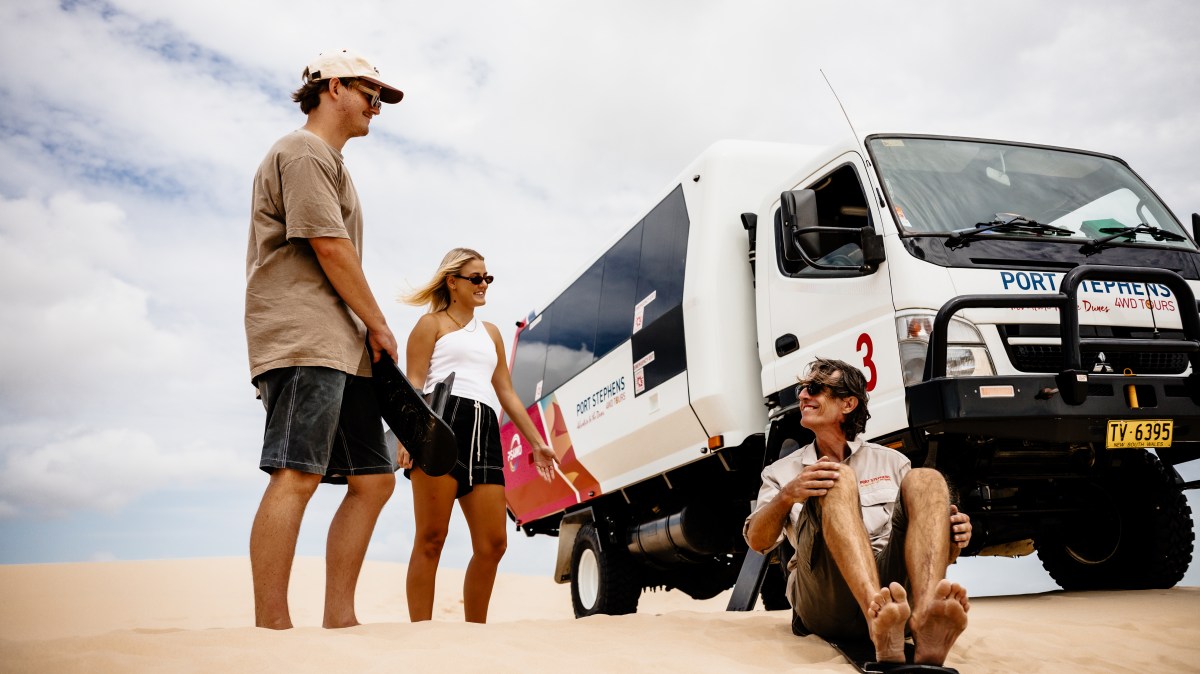 Three people on sand with a tour truck at Port Stephens.