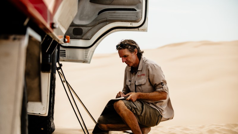 Person kneeling by a truck in a desert, writing on a clipboard.