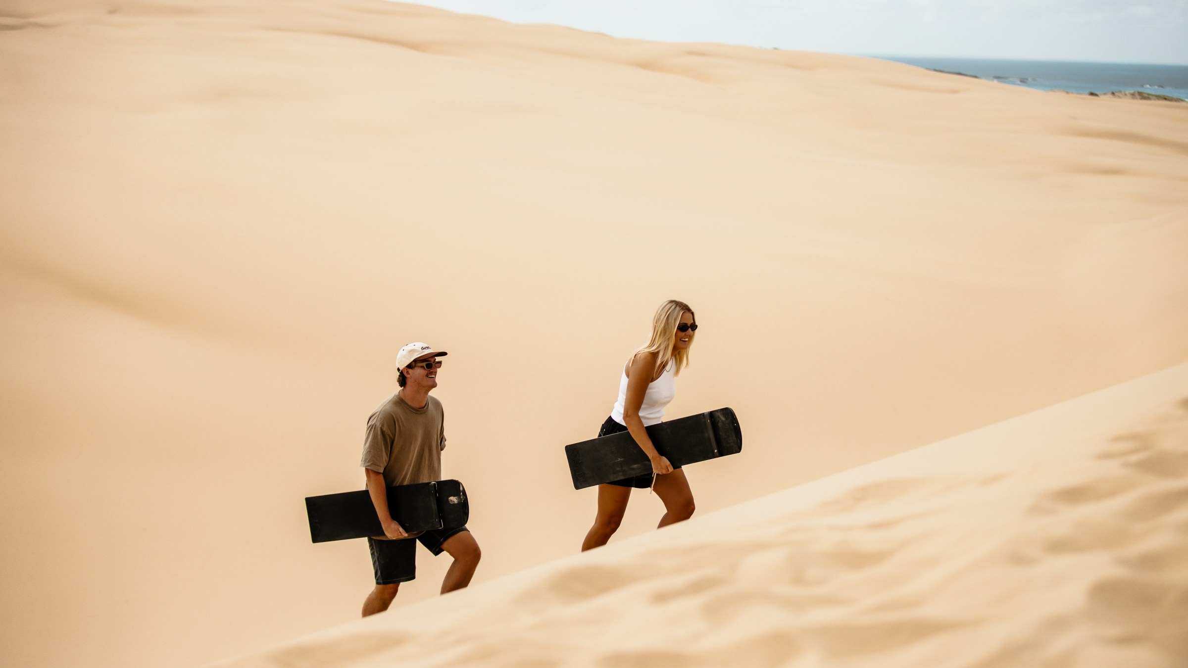 Two people with sandboards walking up a sand dune under a clear sky.