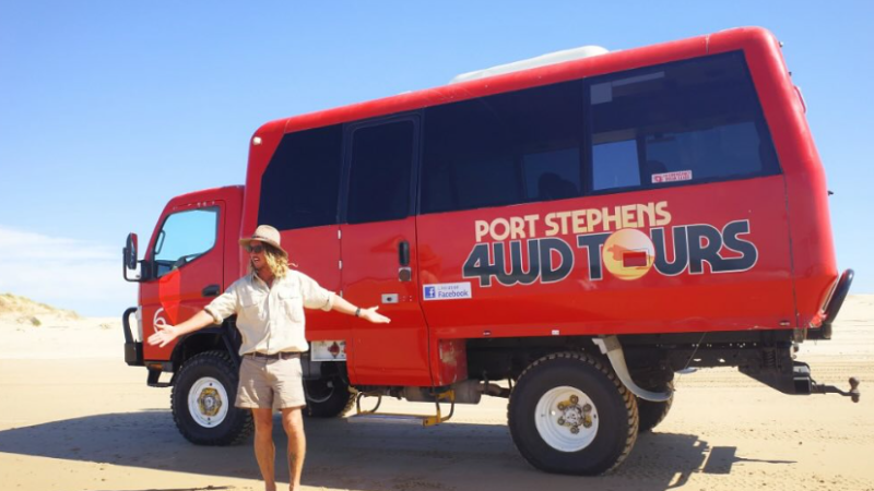 Person in khaki outfit standing next to a red Port Stephens 4WD tour bus on a sandy beach.